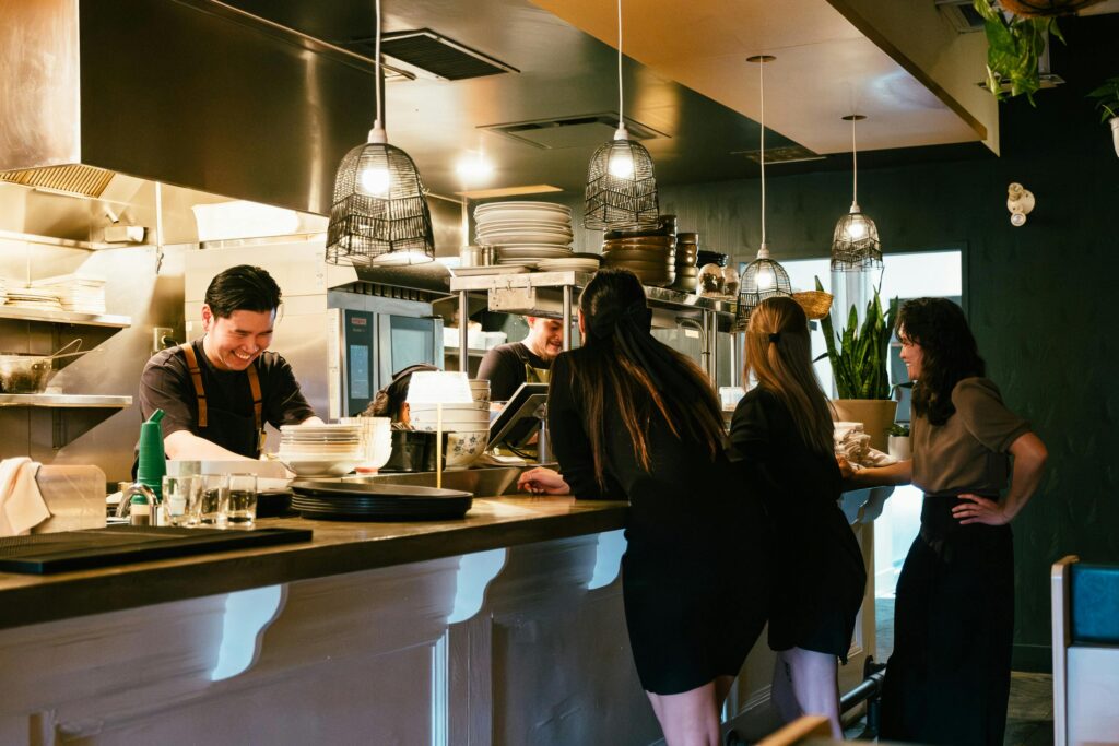 Busy restaurant kitchen with staff preparing meals as patrons watch. Warm lighting and informal dining atmosphere.